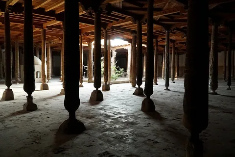 Rows of dark carved timber columns support the flat wooden ceiling of Khiva’s Juma Mosque around a bright roof opening.