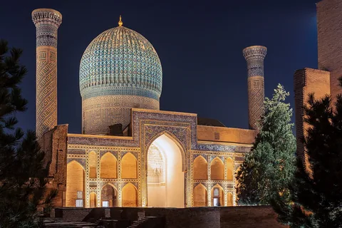 The ribbed turquoise dome, tiled entrance and two minarets of Gur-e Amir illuminated at night.